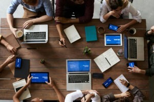 Team sitzt an großem Holztisch, mehrere Laptops, Tablets und Smartphones mit blauen Präsentationsfolien, Notizbücher, Kaffeetassen und Stifte verteilt für Besprechung.